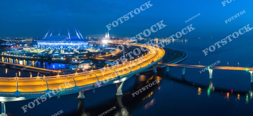 На фотографии изображена магистраль Западного скоростного диаметра (ЗСД) и стадион, расположенные в Санкт-Петербурге.  Яркие огни отражаются в спокойных водах Финского залива, подчёркивая современную архитектуру и инфраструктуру города.